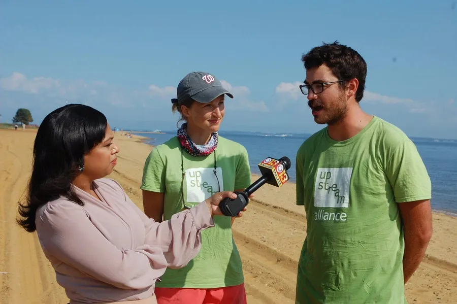 Stephen, in glasses and a green Upstream Alliance shirt, talking to a TV reporter holding a microphone on a Chesapeake beach, with a teammate beside him.