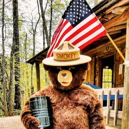 Smokey Bear in a brown fur suit and a campaign hat that reads SMOKEY, holding a clear can in front of a log cabin and an American flag.