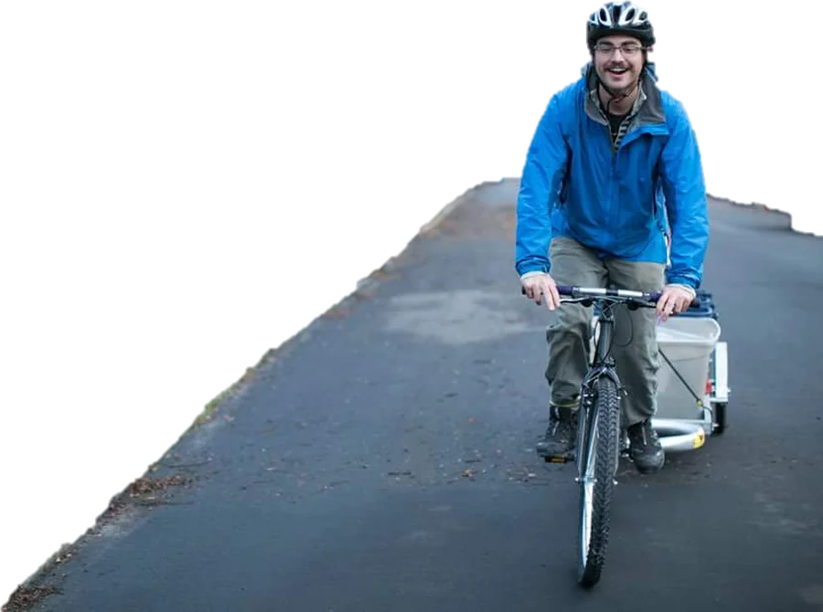 Stephen smiling in a blue rain jacket and bike helmet, riding a mountain bike with a cargo trailer down a paved path.