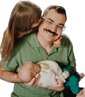 Stephen Eren in a green shirt, smiling and holding his newborn son while his wife leans in to kiss his cheek.